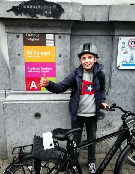 Ein Junge steht mit seinem Fahrrad vor dem Schulschild. Er hebt den Daumen und schaut lächelnd in die Kamera. An seinem Fahrrad ist eine Sensorbox befestigt.